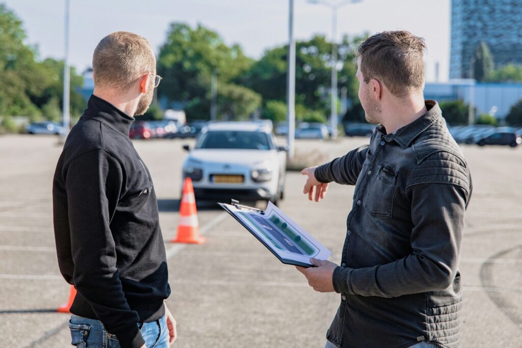 Instructeur geeft rijlessen aan leerling, hij legt iets uit over de auto.